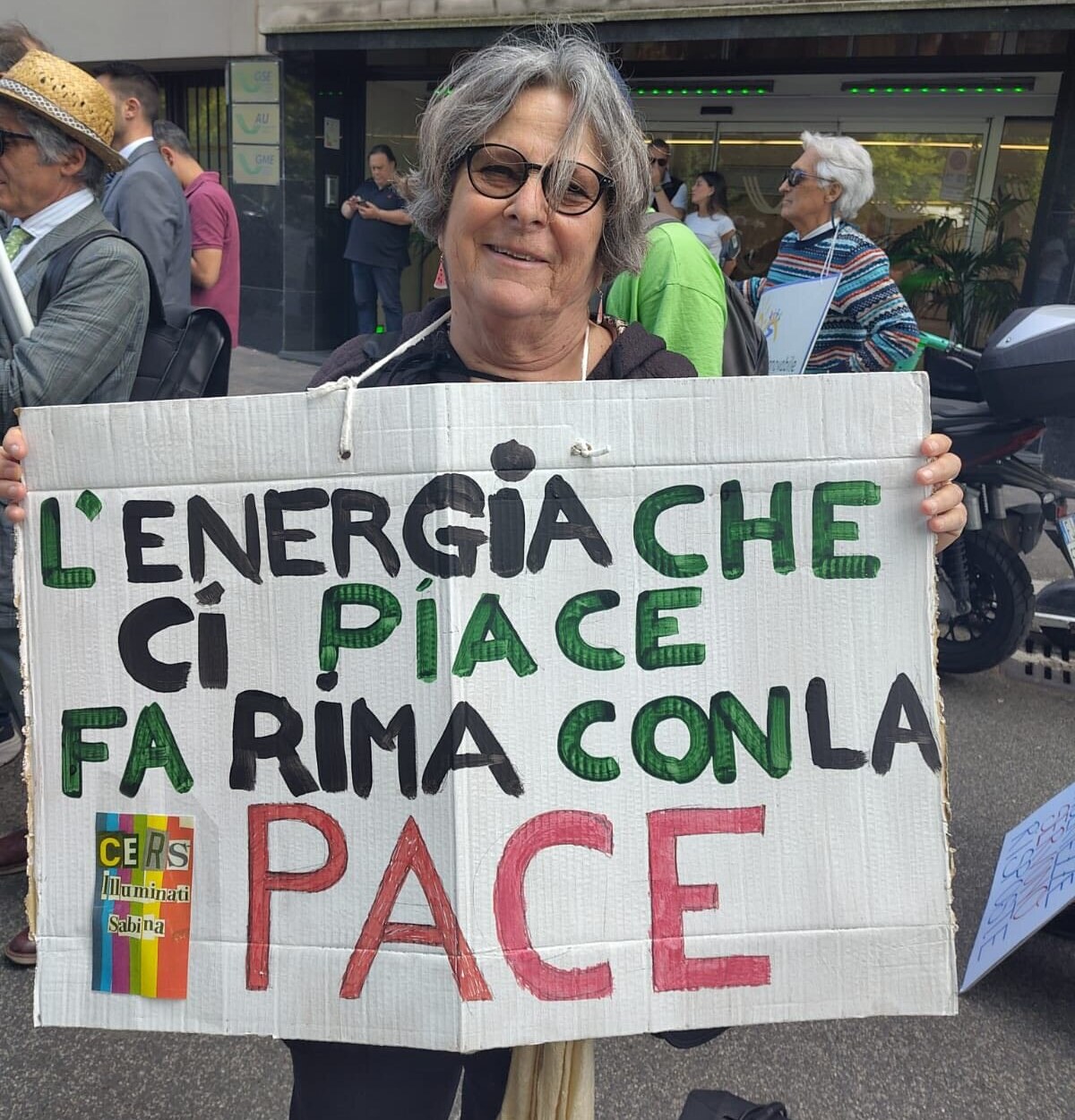 Una manifestante della CERS Illuminati Sabina alla mobilitazione del Movimento Nazionale CER(S) davanti al GSE di Roma il 20 maggio 2026, con striscione 'L'energia che ci piace fa rima con la pace'