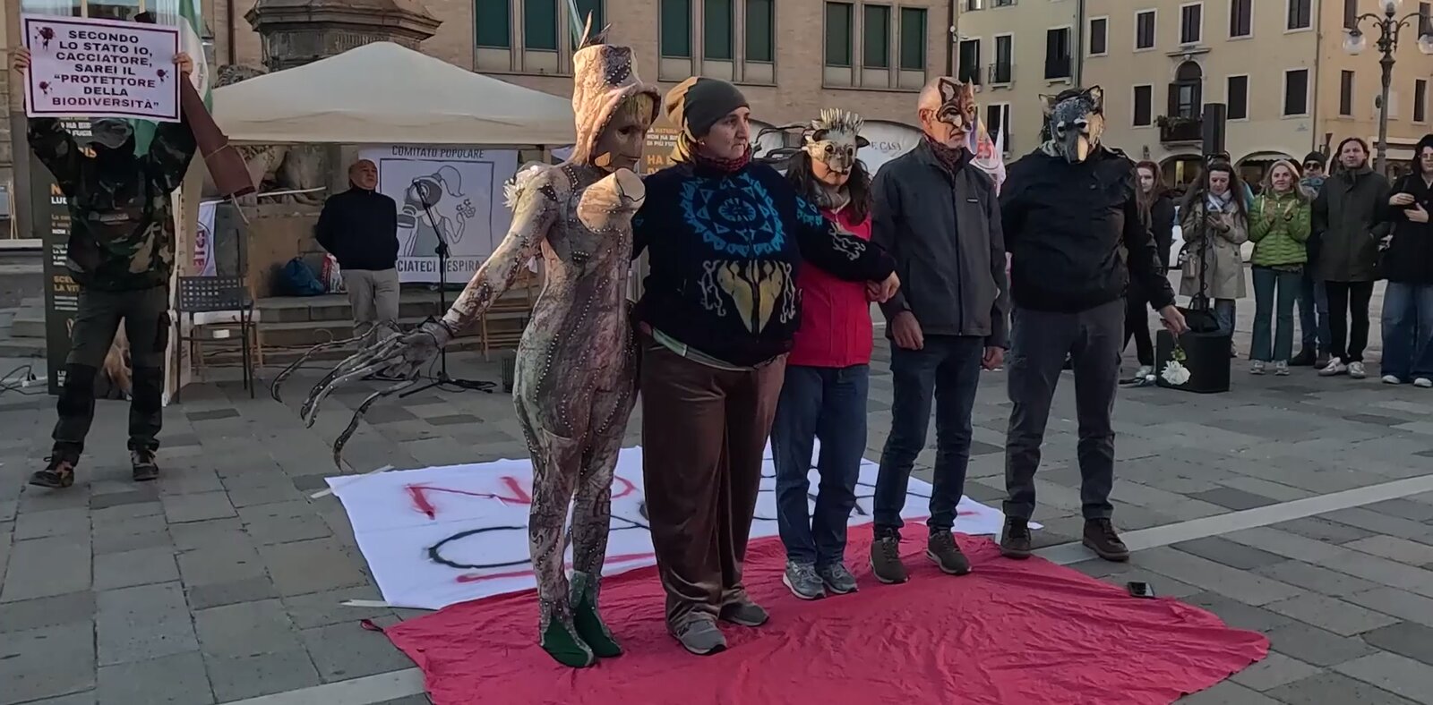 Gli attori della performance in fila sul telo rosso a fine esibizione, con il cervo-albero; a sinistra il cacciatore con il cartello «Secondo lo Stato io, cacciatore, sarei il «protettore della biodiversità»»
